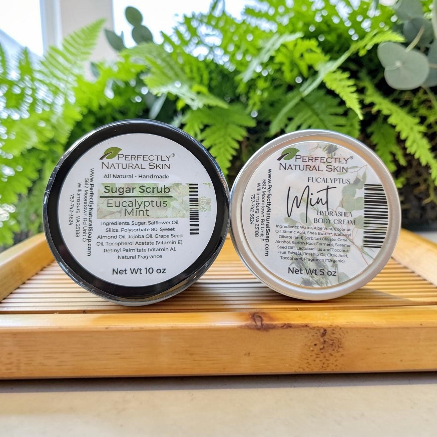 Two small round cosmetic jars from Perfectly Natural Skin, one labeled Sugar Scrub Eucalyptus Mint with a black lid and one labeled Mint with a white lid, displayed on a wooden tray with green plants in the background.