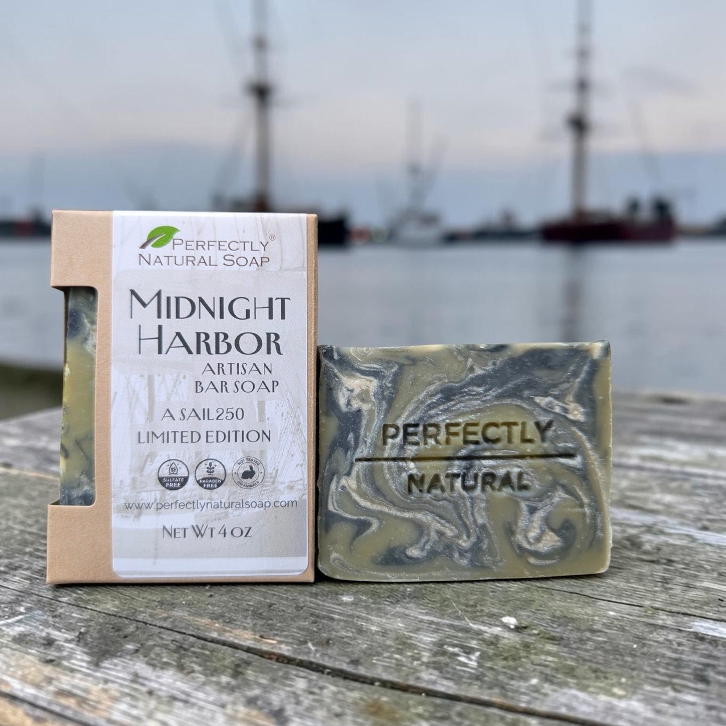 A bar of soap and its packaging box are displayed on a weathered wooden dock with a blurred harbor background. The soap is marbled olive green and gray with 'PERFECTLY NATURAL SOAP' and 'MIDNIGHT HARBOR' printed on it.